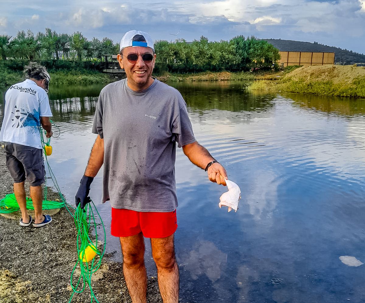 Tevfik Ceyhan smiling at the water's edge in Azmak Stream, holding a piece of chicken in one hand and a rope with a buoy in the other, while a teammate prepares ropes behind him